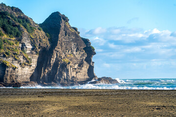 Piha Beach, A striking coastal rock formation bathed in sunlight on a clear day, with the sea and...