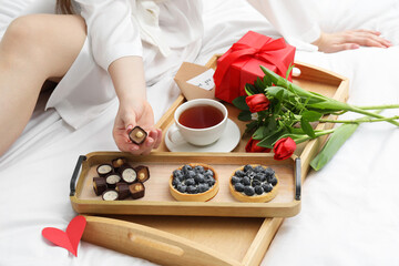 Tasty breakfast served in bed. Woman with desserts, tea, gift box and flowers at home, closeup