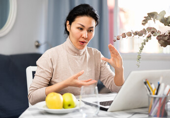 Asian woman sitting at table in apartment and having video call conversation.