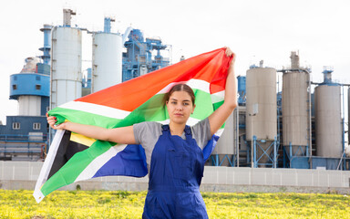 on yellow lawn, near large plant in province,calm girl in overalls of worker stands and holds flag...