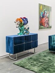 In one corner of the gallery stands a colorful sideboard with a sculpture of stacked stuffed teddy bears in different colors, a beige green patterned carpet covers the floor, and blue velvet sofas lea