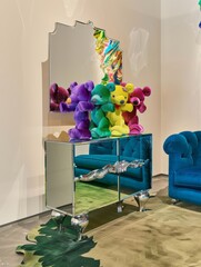 In one corner of the gallery stands a colorful sideboard with a sculpture of stacked stuffed teddy bears in different colors, a beige green patterned carpet covers the floor, and blue velvet sofas lea
