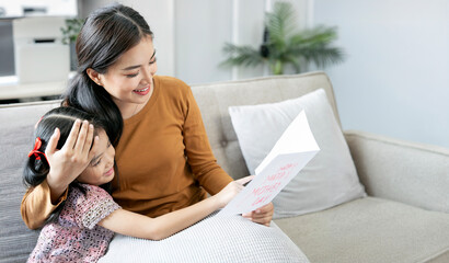 Cheerful asian mother and little daughter reading handmade greeting card  while celebration mothers...
