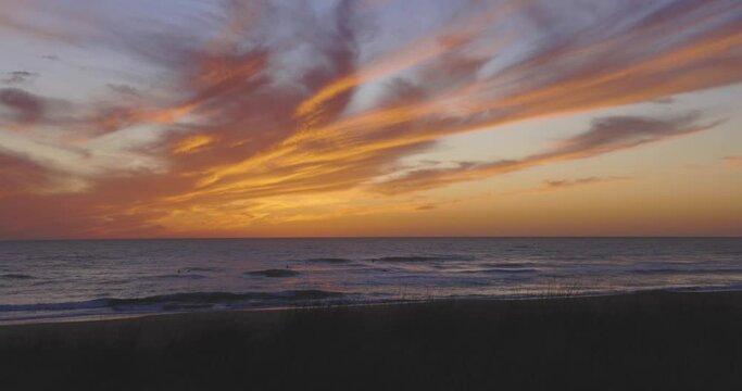 Tropical beach at sunset with people surfing and swimming in ocean with calm waves and dramatic orange sky.