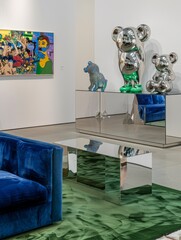 In one corner of the gallery stands a colorful sideboard with a sculpture of stacked stuffed teddy bears in different colors, a beige green patterned carpet covers the floor, and blue velvet sofas lea