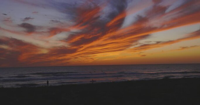  People walking on sand next to ocean with surf boards at dusk. Beach at sunset with dramatic orange and purple sky.