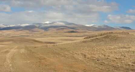 A field road going through the hilly autumn steppe towards high and snow-covered mountain ranges.