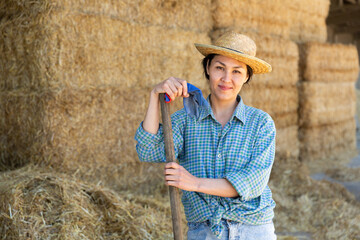 Portrait of positive young asian female farmer standing leaning on pitchfork near straw stack in...