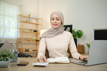 A woman wearing a head scarf sits at a desk with a laptop and a notebook