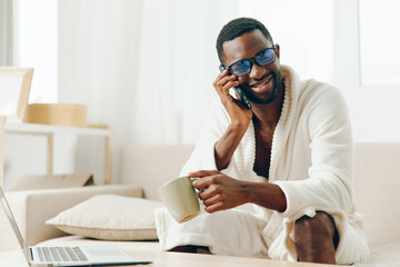 Smiling African American Man Working on Laptop in Cozy Home Office, Talking on Mobile Phone,...