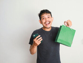 portrait of happy smiling handsome Asian man carrying shopping bags and showing smartphone, online shopping concept