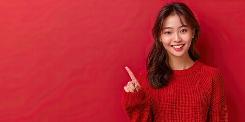 Smiling young asian woman with long wavy hair pointing finger while wearing red knitted sweater against bright red background