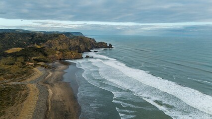 A dramatic aerial view of coastal cliffs with rolling waves crashing against the rocky shore, under a cloudy sky  - Bethells Beach