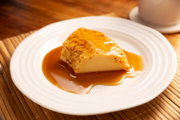 Slice of homemade flan dessert, also known as creme caramel, caramel custard pudding or egg custard, served on white plate on wooden table.