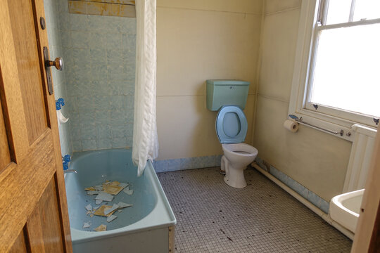 An old and dusty hotel bathroom and toilet with outdated interiors; wall tiles collapse and fall into the bathtub. Concept of a renovation project, abandoned house, or rundown accommodation.