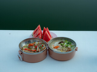 Breakfast set watermelon fruit, pork porridge, pan-fried eggs arranged in containers on white table by the lake.
