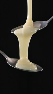 White sweet condensed milk sauce flowing from metal spoon on black background. Food close up, slow motion, vertical video 4K