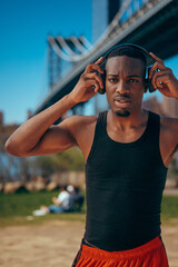 Young man in sportswear adjusting headphones near a bridge in a park