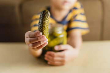 A little boy is holding a cucumber pickle and a glass jar of pickle, with the background of a...