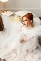 Graceful young woman with fiery red hair and flawless makeup in a stunning white bridal gown. She...
