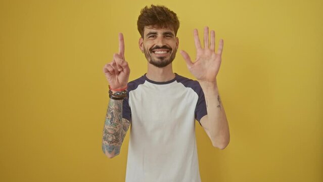 Joyous young hispanic man, smiling and confidently pointing up with six fingers, standing over isolated yellow background, happily showing the number six