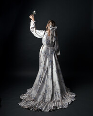 portrait of beautiful blonde female model wearing romantic historical gown white bridal floral pattern. standing pose running away from camera with flowing silk skirt. isolated dark studio background.