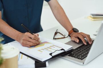 Cropped shot professional businessman analyzing graph paper and using laptop at desk