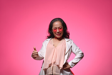 Confident cool senior woman in trendy glasses showing thumbs up gesture over pink background