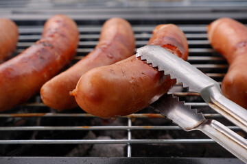 Close-up of flipping grilled sausages with metal tongues. Chef preparing delicious food for summer...