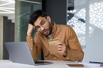 Man looking concerned while holding credit card and using laptop at work
