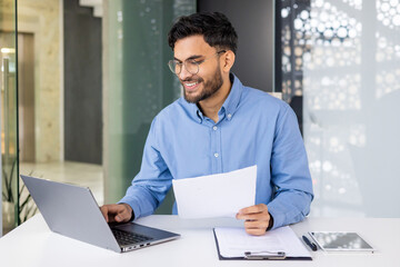 Smiling businessman working on laptop and holding paper while sitting in modern office