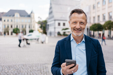 A confident mature businessman in a blue suit smiling while holding a smartphone in an urban...