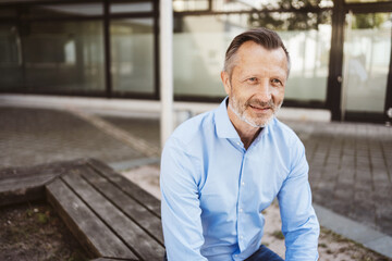 Mature man sitting outdoors wearing a blue shirt, with a modern office background. He looks...