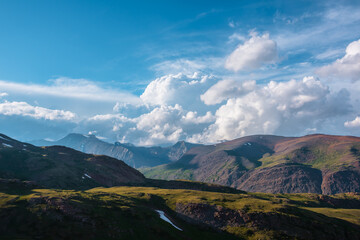 Awesome alpine landscape with sunlit green hills and multicolor mountains under huge lush clouds in...