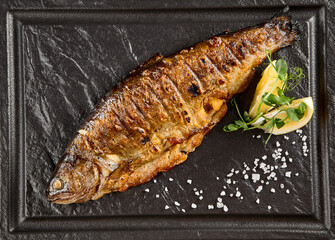 Grilled rainbow trout with a lemon wedge and fresh herbs on a dark stone plate