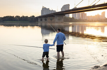 Dad and son fish together, standing with fishing rods in the river water at sunset. father's day...