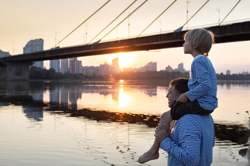 Portrait of father and son in identical long sleeves watching sunset by the river. child boy sits...