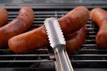 Metallic tongues holding grilled sausage in front of bbq grill.