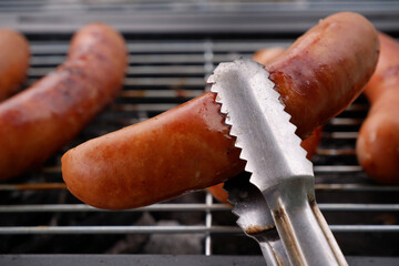 Metal tongues holding grilled sausage in front of bbq grill close up