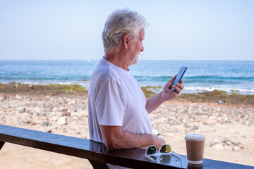 Portrait of bearded senior retired man outdoors at the seaside beach talking on mobile phone enjoying free time and relaxed vacation. Horizon over water