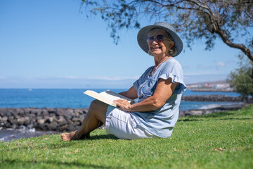 Smiling senior woman in straw hat sitting barefoot in meadow face the sea reading a book, relaxed senior lady enjoying free time in summer vacation or retirement