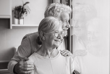 Black and white portrait of bonding senior retired couple standing near window, looking in distance, recollecting good memories or planning common future, enjoying peaceful moment together at home