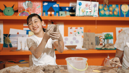 Asian highschool student modeling clay on table with mud and bottle of water at pottery workshop....