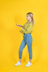 Impressed woman using laptop, full body side view young blonde caucasian impressed woman using laptop. Standing over yellow studio background. Looking notebook screen shocked face expression. 
