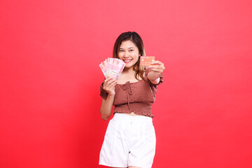 Asian woman smiling, holding rupiah money and debit credit card in front, wearing a brown blouse,...