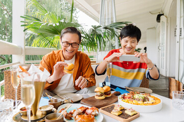 Happy Asian Senior mature married couple Enjoying Healthy Lunch Outdoors on a open air Sunny Terrace. on vacation in a sunny summer day, while the woman is taking photos with cell phone