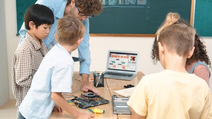 Cute energetic children working on electronic board while teacher using tablet analyze data. Group of diverse student fixing main board and learning about generated AI and programing system. Pedagogy.