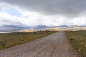 A gravel road through autumn fields leading to a ridge of mountains covered with dense clouds.