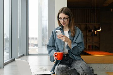 Young female student holding a cup and using a smartphone near the window.