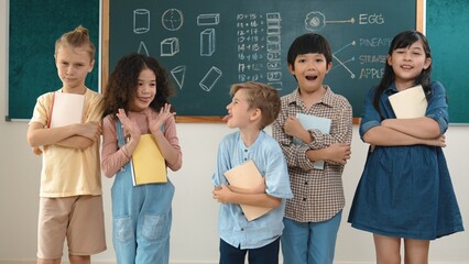 Multicultural elementary student in casual cloth looking at camera while standing at classroom. Group of cute diverse children hold notebook and smiling to camera at blackboard in classroom. Pedagogy.
